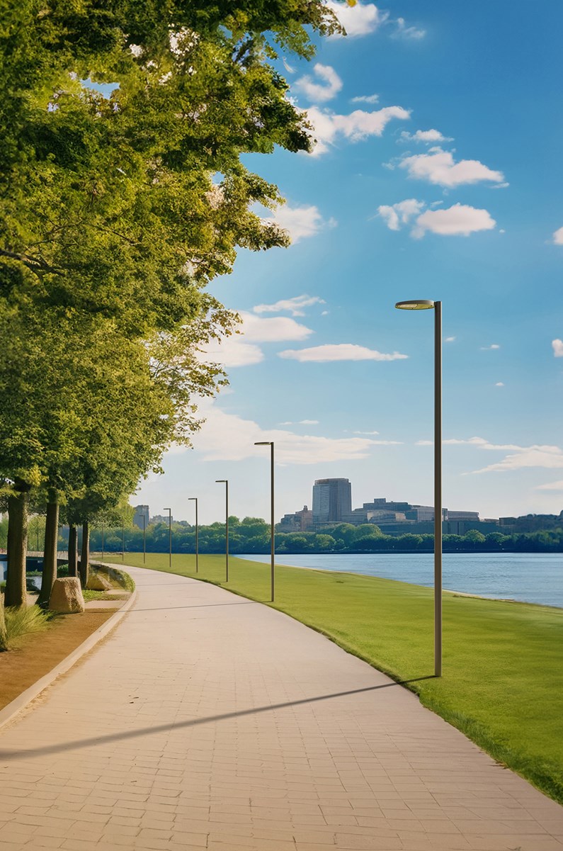 A scenic riverside pathway lined with modern AEC Illuminazione NOOS luminaires mounted on sleek columns. The pathway curves gently alongside lush green grass and trees, with a cityscape visible in the background under a bright blue sky with scattered clouds.